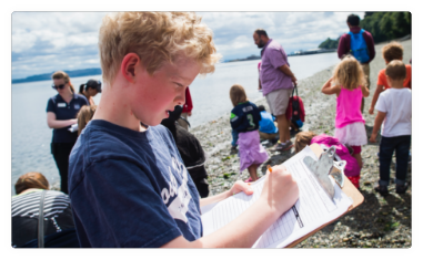A young teenage fills out a form while standing on a beach amongst a crowd of all ages.