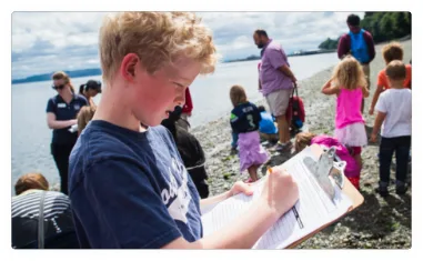 A young teenage fills out a form while standing on a beach amongst a crowd of all ages.
